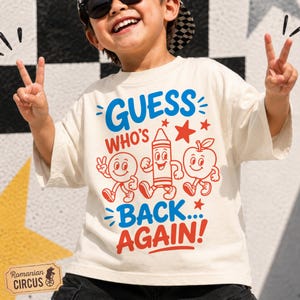 Może przedstawiać: Kremowy t-shirt z krótkim rękawem z nadrukiem graficznym zawierającym napis "GUESS WHO'S BACK... AGAIN!" w kolorze niebieskim i czerwonym. Wzór zawiera rysunkowe ilustracje uśmiechniętej buzi, kredki oraz jabłka w czerwonym kolorze.