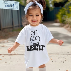 Puede incluir: Una camiseta blanca con una mano de dibujos animados negra haciendo un signo de paz y la palabra "LEGIT" en una fuente retro.