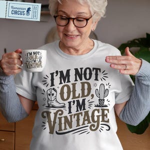 Könnte beinhalten: Weißes T-Shirt mit dem Text "I'M NOT OLD, I'M VINTAGE" in Schwarz und Braun. Das Design zeigt einen Cowboy-Stiefel und einen Kaktus. Eine weiße Tasse mit dem gleichen Text wird gehalten.
