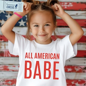 May include: A white t-shirt with the red text "ALL AMERICAN BABE" is worn by a young girl. The background features a blurred American flag. A small ticket with the text "Romanian Circus" is visible.