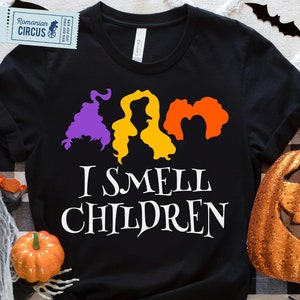 May include: Black t-shirt with a Halloween theme. The shirt features three witch silhouettes in purple, yellow, and orange, with the text "I SMELL CHILDREN" in white. A pumpkin and other Halloween decorations are in the background.