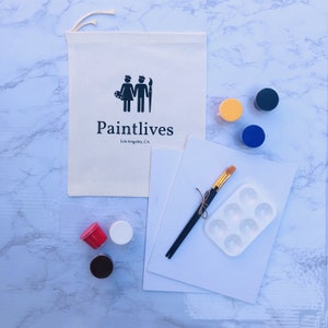 May include: A paint and sip kit with a white canvas, six paint pots, two paintbrushes, and a white plastic paint palette. The kit also includes a white canvas bag with the text "Paintlives Los Angeles, CA" and a black silhouette of two people painting.