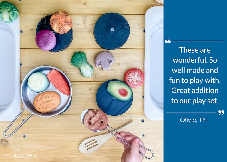 May include: A set of wooden play food items, including a loaf of bread, a roll, a carrot, a cucumber, an avocado, a mushroom, a tomato, and a green leafy vegetable. The food items are arranged on a wooden surface with a white sink and a stainless steel pan.