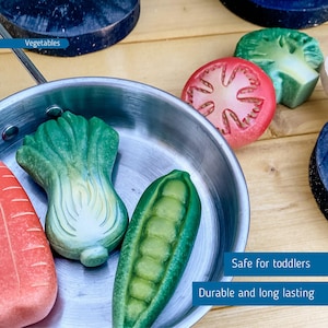 May include: A silver frying pan with a green lettuce, a green pea pod, and a red tomato slice. The vegetables are made of wood and are part of a toy food set. The text "Safe for toddlers" and "Durable and long lasting" is visible in the image.