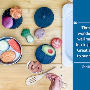 May include: A set of wooden play food items, including a loaf of bread, a roll, a carrot, a cucumber, an avocado, a mushroom, a tomato, and a green leafy vegetable. The food items are arranged on a wooden surface with a white sink and a stainless steel pan.