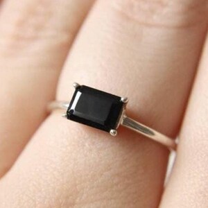 May include: A silver ring featuring a rectangular, black gemstone. The ring's band is thin and metallic, with the dark stone set in a four-prong setting. The ring is shown on a finger.
