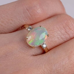 May include: A gold ring featuring an oval opal gemstone with iridescent colors. The ring is accented with two small, round, clear gemstones on either side of the opal. The ring is set on a light skin tone.