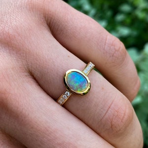 May include: A gold ring featuring an oval opal gemstone with blue and green hues. The ring has a gold band with small, clear gemstones set on each side of the opal. The ring is shown on a person's finger.