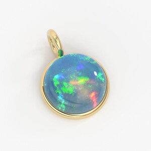 May include: A round pendant featuring a polished cabochon opal set in a smooth yellow gold bezel. The opal displays a dark blue base color with vibrant flashes of green, red, and blue light. A small gold bail is attached to the top of the setting.