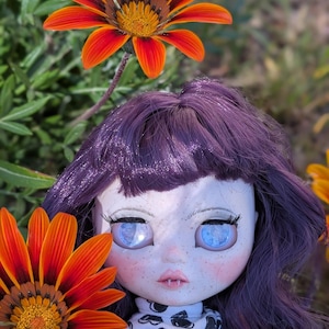 May include: A doll with purple hair and large blue eyes, adorned with freckles, is surrounded by vibrant orange and yellow flowers. The doll wears a black and white patterned turtleneck.