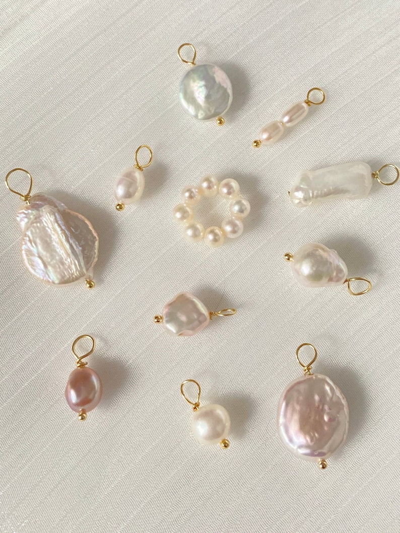 K&ouml;nnte beinhalten: Sortiment an wei&szlig;en und rosafarbenen Perlenanh&auml;ngern mit goldenen Akzenten. Die Anh&auml;nger haben verschiedene Formen, darunter rund, oval und unregelm&auml;&szlig;ig. Einige Anh&auml;nger sind zu einem Ring zusammengef&uuml;gt.
