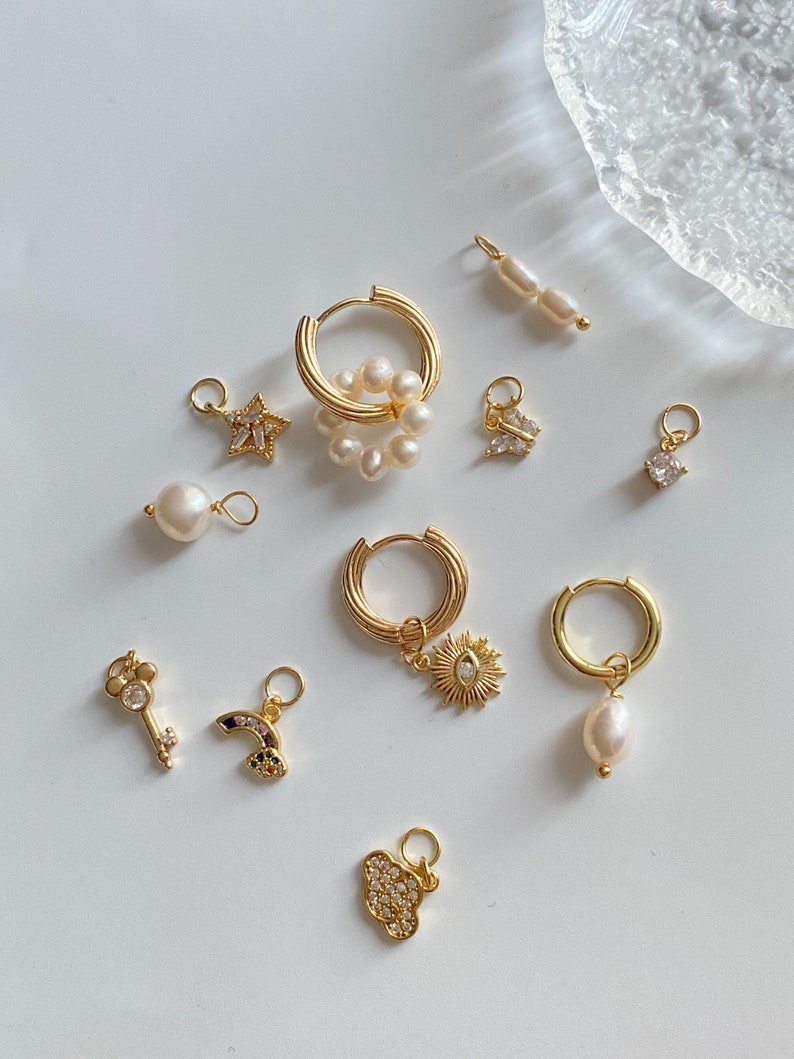 K&ouml;nnte beinhalten: Eine Sammlung goldener Creolen mit verschiedenen Anh&auml;ngern, darunter eine Perle, ein Stern, ein Schmetterling, eine Sonne, ein Regenbogen, ein Schl&uuml;ssel, eine Wolke und ein Quadrat. Die Anh&auml;nger sind aus Gold und haben kleine Kristalle oder Perlen.