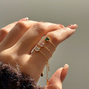 Peut inclure: Une bague en chaîne dorée avec trois petites pierres précieuses. Les pierres précieuses sont vertes, roses et claires.