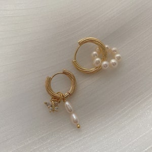 K&ouml;nnte beinhalten: Ein Paar goldene Creolen mit Perlenakzenten. Ein Ohrring hat einen kleinen Schmetterlingsanh&auml;nger mit klaren Strasssteinen, w&auml;hrend der andere Ohrring einen Perlenschwarm hat, der vom Reifen h&auml;ngt.