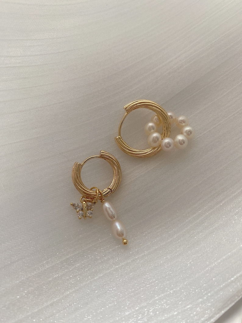 K&ouml;nnte beinhalten: Ein Paar goldene Creolen mit Perlenakzenten. Ein Ohrring hat einen kleinen Schmetterlingsanh&auml;nger mit klaren Strasssteinen, w&auml;hrend der andere Ohrring einen Perlenschwarm hat, der vom Reifen h&auml;ngt.
