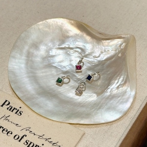 May include: Four silver charms with different coloured gemstones. The charms are on a white mother of pearl surface. The charms are a red square, a green square, a blue square, and a silver padlock.
