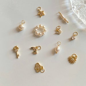 K&ouml;nnte beinhalten: Eine Sammlung von goldenen Charms mit verschiedenen Designs, darunter ein Stern, ein Schmetterling, ein Schl&uuml;ssel, ein Regenbogen, eine Wolke, eine Sonne, eine Perle und ein Perlring. Die Charms sind auf einer wei&szlig;en Oberfl&auml;che angeordnet.