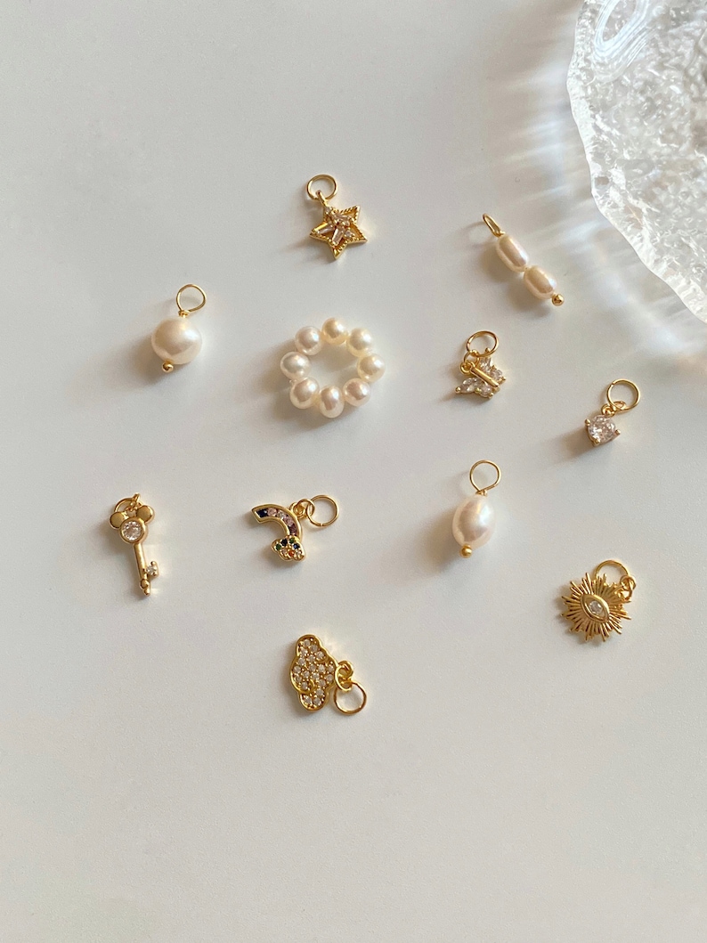 K&ouml;nnte beinhalten: Eine Sammlung von goldenen Charms mit verschiedenen Designs, darunter ein Stern, ein Schmetterling, ein Schl&uuml;ssel, ein Regenbogen, eine Wolke, eine Sonne, eine Perle und ein Perlring. Die Charms sind auf einer wei&szlig;en Oberfl&auml;che angeordnet.