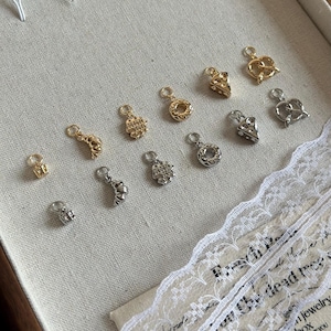 Peut inclure: Une collection de 12 breloques dorées et argentées avec différents motifs, dont un bretzel, un cœur, une étoile et un croissant de lune. Les breloques sont disposées sur une surface blanche avec un recouvrement de dentelle.