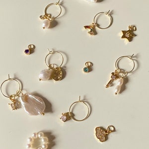 K&ouml;nnte beinhalten: Eine Sammlung von goldenen Creolen mit verschiedenen Anh&auml;ngern, darunter eine Perle, ein Schmetterling, ein Stern, ein Schl&uuml;ssel, ein Regenbogen, eine Wolke und eine M&uuml;nze. Die Anh&auml;nger sind mit kleinen Sprungringen an den Creolen befestigt.
