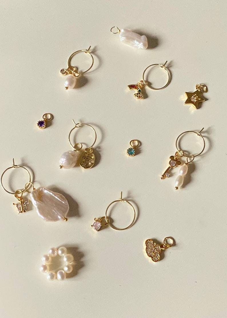 K&ouml;nnte beinhalten: Eine Sammlung von goldenen Creolen mit verschiedenen Anh&auml;ngern, darunter eine Perle, ein Schmetterling, ein Stern, ein Schl&uuml;ssel, ein Regenbogen, eine Wolke und eine M&uuml;nze. Die Anh&auml;nger sind mit kleinen Sprungringen an den Creolen befestigt.