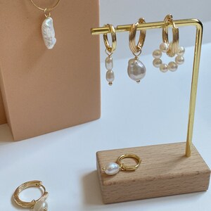 K&ouml;nnte beinhalten: Ein Set goldener Creolen mit Perlenanh&auml;ngern. Die Ohrringe werden auf einem Holzst&auml;nder pr&auml;sentiert. Es gibt auch eine goldene Halskette mit einem Perlenanh&auml;nger.