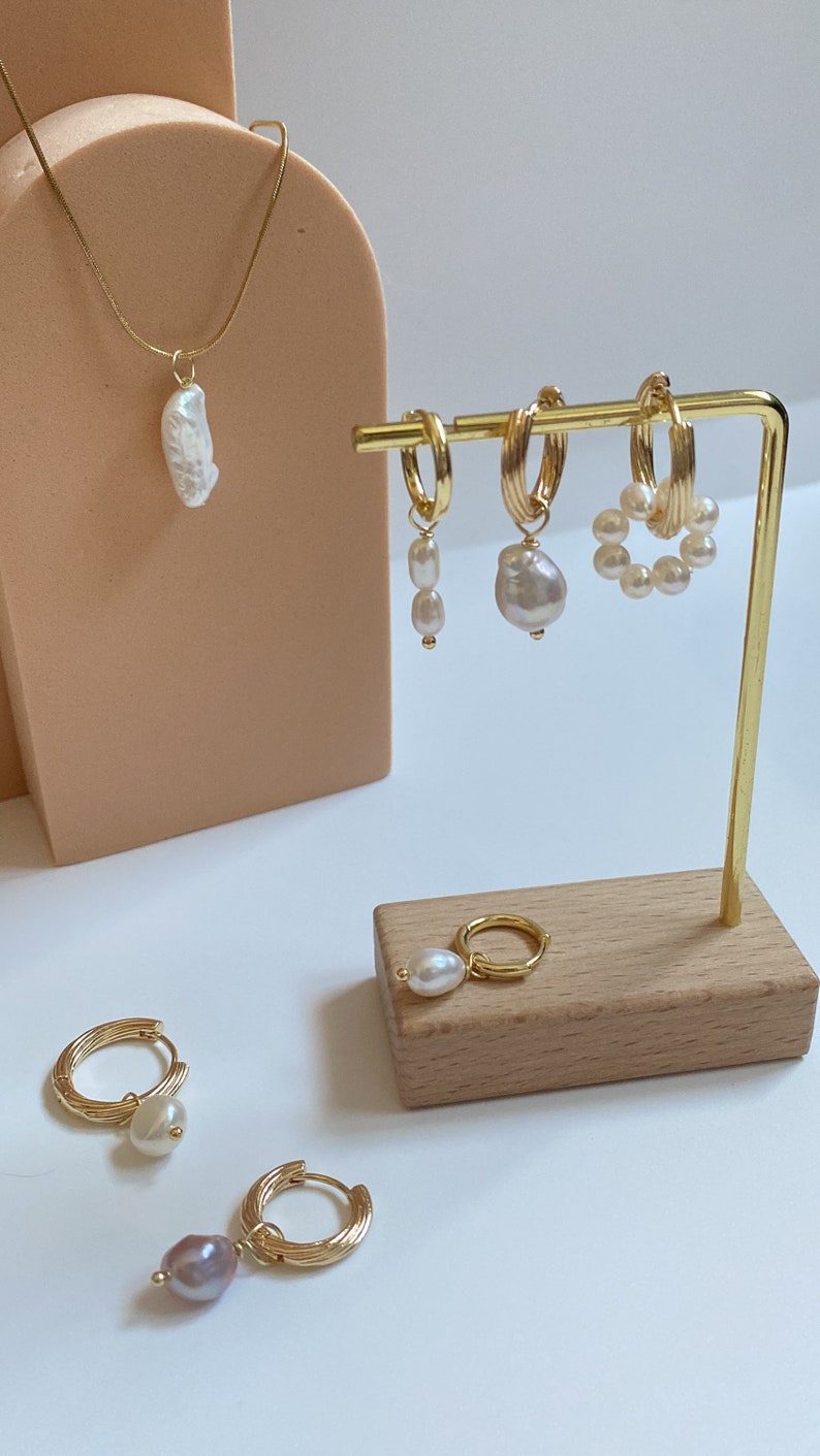K&ouml;nnte beinhalten: Ein Set goldener Creolen mit Perlenanh&auml;ngern. Die Ohrringe werden auf einem Holzst&auml;nder pr&auml;sentiert. Es gibt auch eine goldene Halskette mit einem Perlenanh&auml;nger.