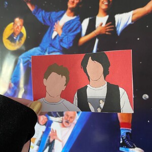 May include: A red card with a cartoon illustration of two men. The men are wearing casual clothing and have their faces obscured. The card is held in front of a movie poster for "Bill & Ted's Excellent Adventure".