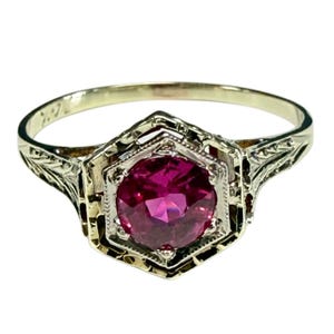 May include: A gold ring featuring a round, faceted, deep pink gemstone set within a hexagonal frame. The metal band displays intricate, engraved leaf-like patterns along the sides, leading up to the central setting with a beaded edge detail.