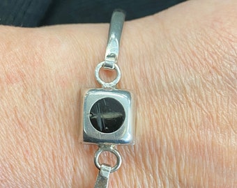 May include: A silver bangle bracelet with a square charm featuring a black circle in the center.