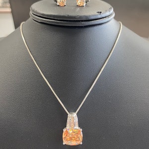 May include: A silver necklace with a pendant featuring a large, square-shaped, orange gemstone surrounded by smaller, clear gemstones. The necklace is displayed on a black mannequin. Matching earrings with orange gemstones are also shown.