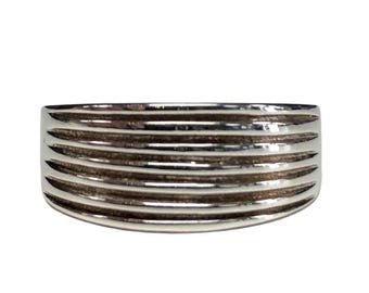 May include: A silver-toned ring with a ribbed design. The ring features multiple horizontal grooves across its surface, creating a textured appearance. The ring is a wide band style, with a smooth, polished finish.
