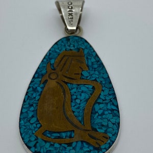 May include: A silver pendant with a turquoise inlay depicting a stylized figure. The pendant is oval shaped and has a silver bail. The back of the pendant is stamped "MEXICO".
