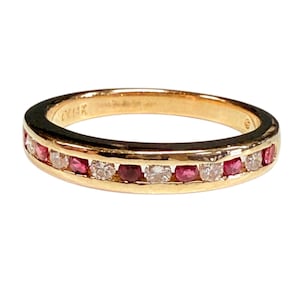 May include: A gold band ring featuring a channel setting with alternating round-cut red rubies and clear white diamonds. The interior of the band is clearly stamped with the text 14K, indicating the gold purity of this jewelry piece.