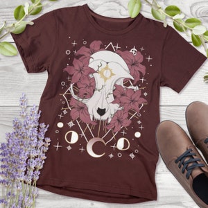 May include: A maroon t-shirt featuring a graphic print of a white animal skull surrounded by pink flowers. The skull has a gold sun symbol in its forehead. The graphic is surrounded by a geometric pattern with gold accents and white stars.