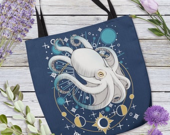 May include: Navy blue tote bag featuring a detailed illustration of a white octopus with teal accents. The design includes celestial elements like stars, moons, and planets. The bag has black handles and is set against a wooden background.