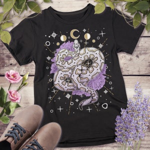 May include: Black t-shirt with a graphic of a white snake with a geometric pattern, surrounded by purple roses and gold stars and moons.