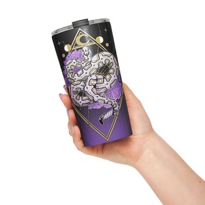 May include: A stainless steel travel tumbler with a black lid and a purple and black body. The tumbler features a white and purple snake design with gold geometric shapes and purple flowers. The tumbler is held in a hand.