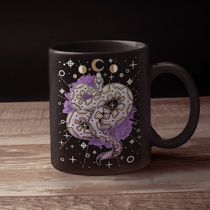 May include: Black ceramic mug with a white snake design with purple flowers and gold celestial symbols. The snake is coiled around the flowers and has a geometric pattern.