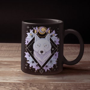 May include: A black ceramic mug featuring a wolf illustration. The wolf is surrounded by flowers and moon phases, set within a geometric diamond shape. The design incorporates shades of purple, white, and gold.