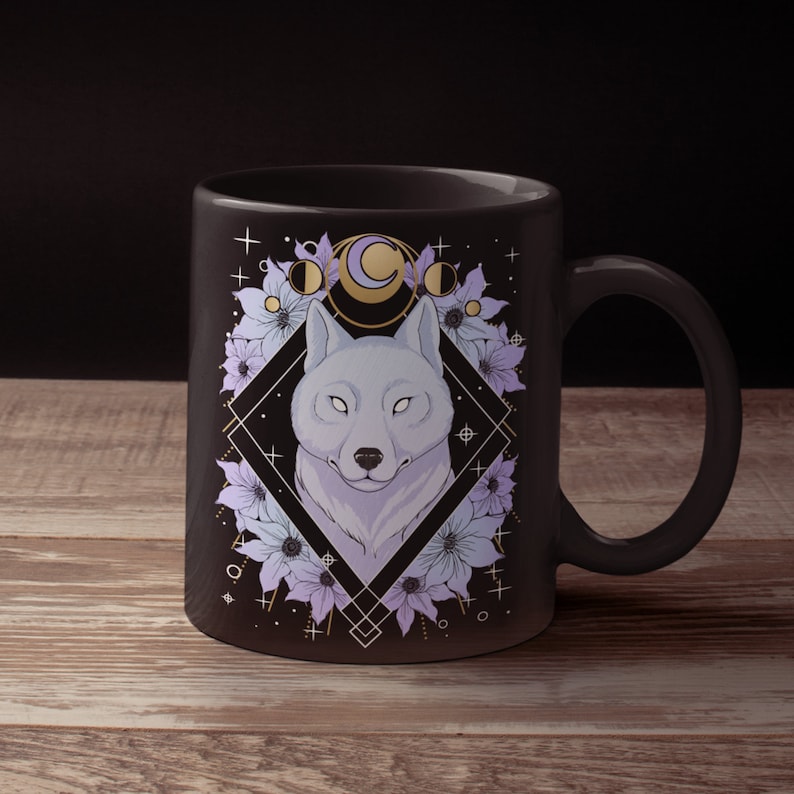 May include: A black ceramic mug featuring a wolf illustration. The wolf is surrounded by flowers and moon phases, set within a geometric diamond shape. The design incorporates shades of purple, white, and gold.
