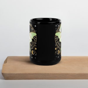 Puede incluir: Taza de cer&aacute;mica negra con un dise&ntilde;o de polilla verde y dorada. La polilla est&aacute; rodeada de un dise&ntilde;o circular con estrellas y lunas.