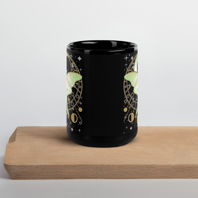 Puede incluir: Taza de cer&aacute;mica negra con un dise&ntilde;o de polilla verde y dorada. La polilla est&aacute; rodeada de un dise&ntilde;o circular con estrellas y lunas.