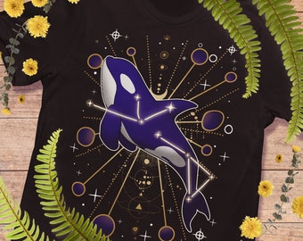 May include: Black t-shirt with a purple and white orca whale design, incorporating a constellation pattern. The design includes stars and circles. The shirt is framed by yellow flowers and green ferns, creating a natural aesthetic.