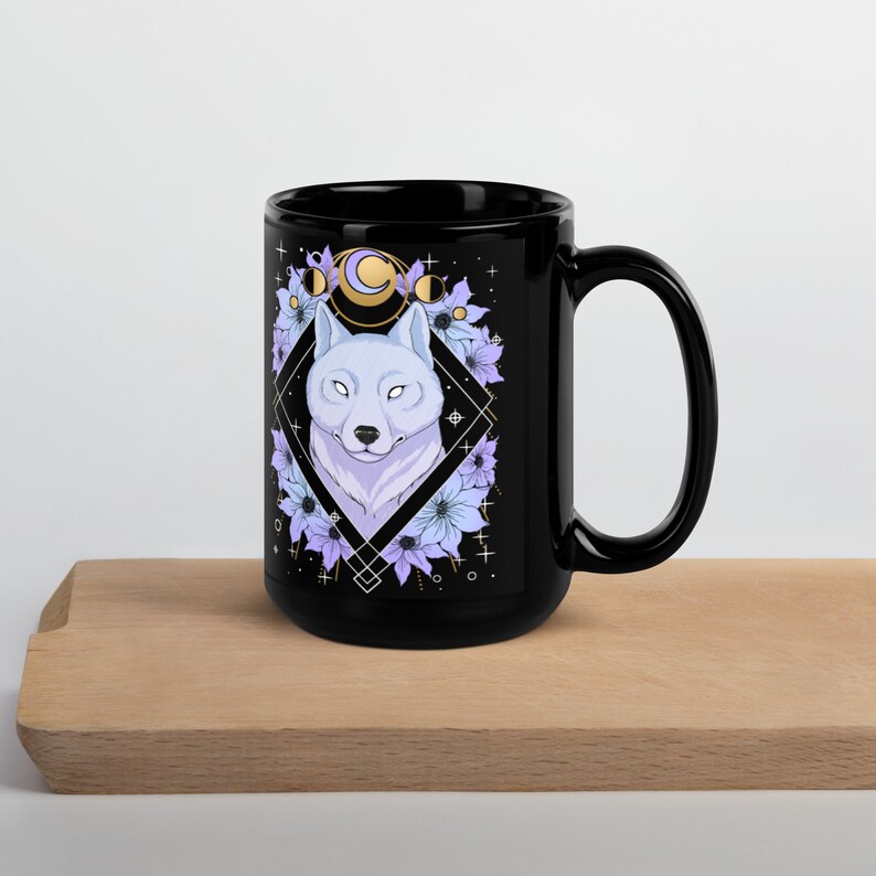 May include: Black ceramic mug featuring a white wolf illustration within a diamond frame, surrounded by purple and blue flowers and celestial elements. The mug has a large handle and is suitable for hot or cold beverages.