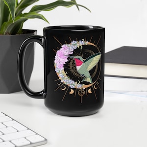 May include: Black ceramic mug featuring a hummingbird design. The design includes a hummingbird with green and red feathers, a crescent moon, and floral accents in pink and blue. The mug is on a white surface.