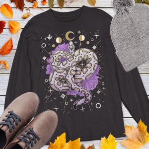 Puede incluir: Camiseta negra de manga larga con un diseño detallado de una serpiente, fases lunares y elementos florales. La serpiente es de color blanco, dorado y morado. Un gorro gris con pompón y zapatos marrones también están en la imagen.