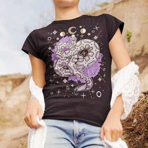 May include: Black t-shirt featuring a white snake design, purple flowers, and gold celestial symbols. The snake is curled up with its head facing the left side of the design.
