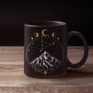 May include: A black ceramic mug featuring a gold illustration of a mountain range, forest, stars, and moon phases. The design is enclosed in a circular frame. The mug has a curved handle and sits on a wooden surface.