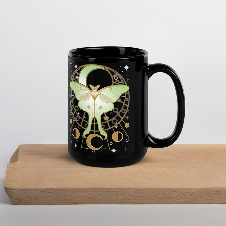Puede incluir: Taza de cer&aacute;mica negra con un dise&ntilde;o de polilla luna verde y dorada. La polilla est&aacute; rodeada por una luna creciente y estrellas.